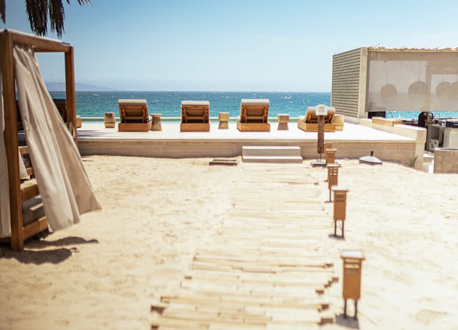 A sandy beach with wooden loungers facing the ocean and a pathway leading to a modern structure.