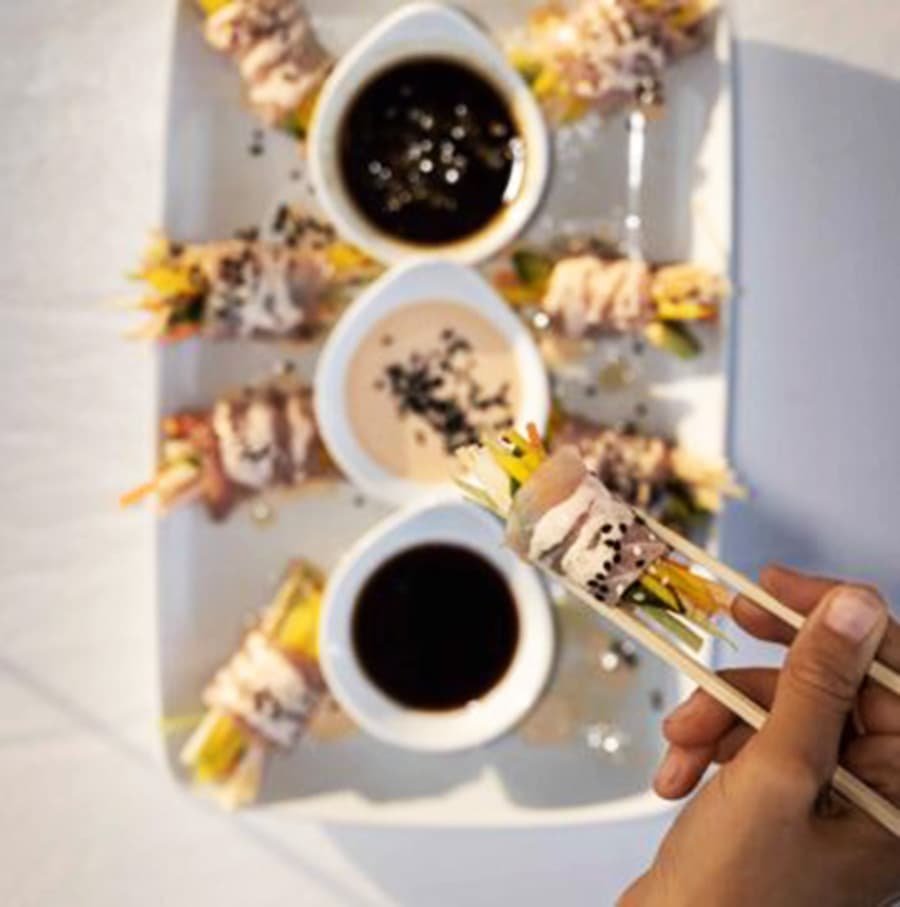 A hand uses chopsticks to pick up a colorful sushi roll beside dipping sauces on a plate.