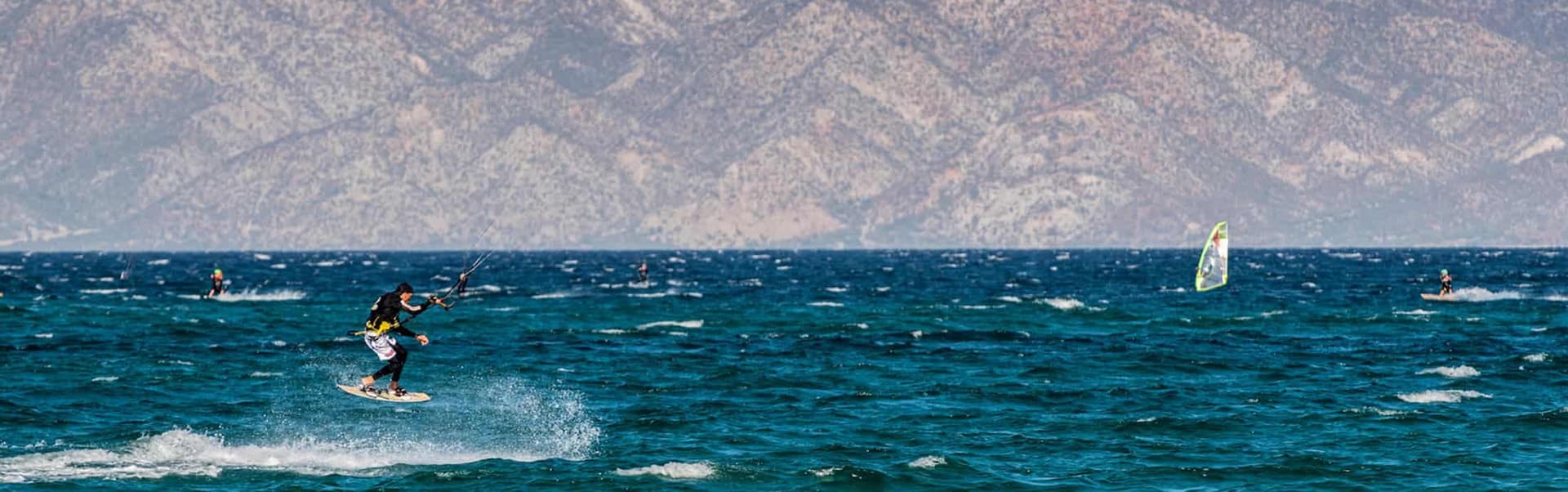A kiteboarder jumps over waves with mountains in the background.