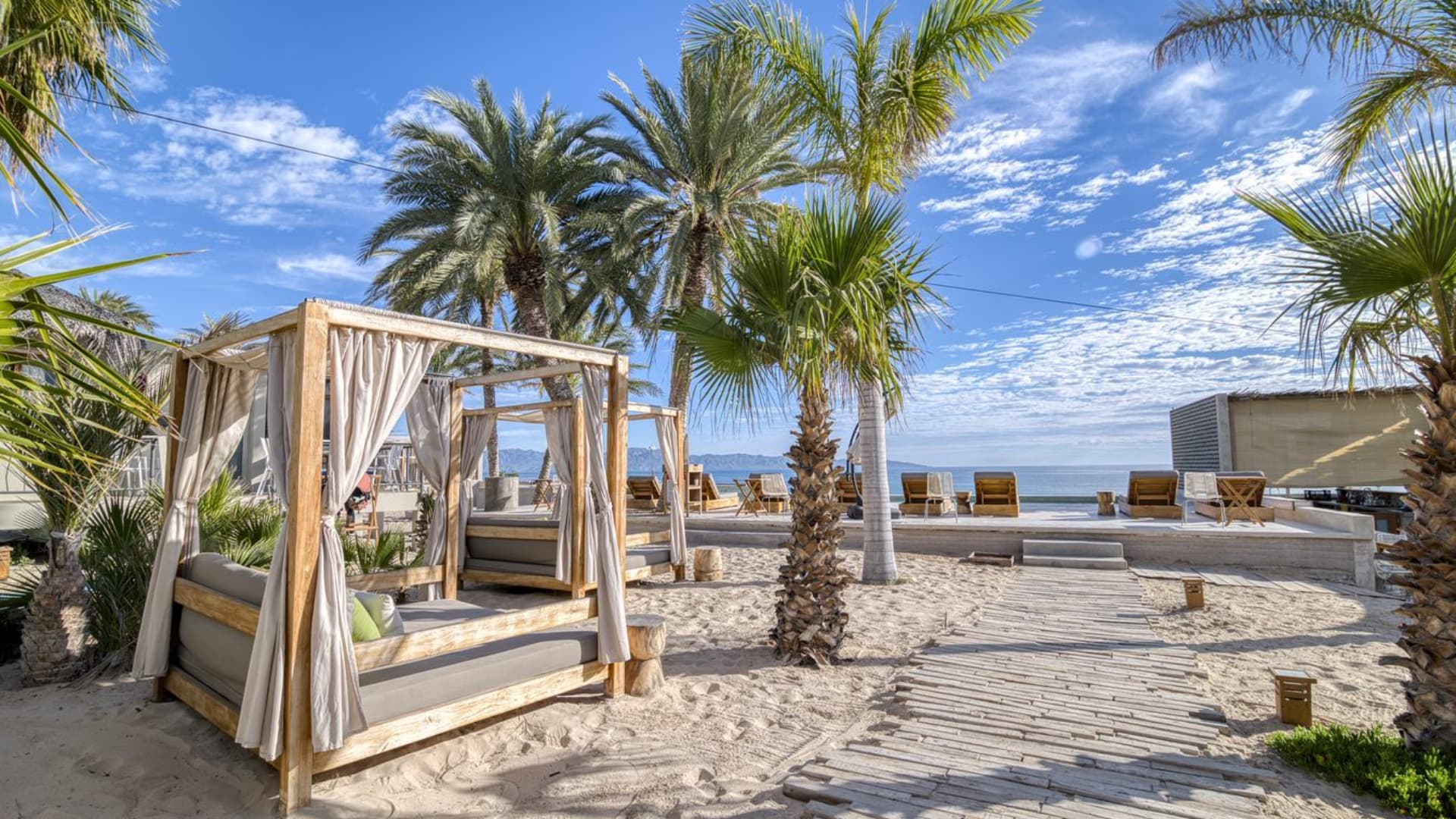Tropical beach lounge area with canopy beds, palm trees, and a clear blue sky.