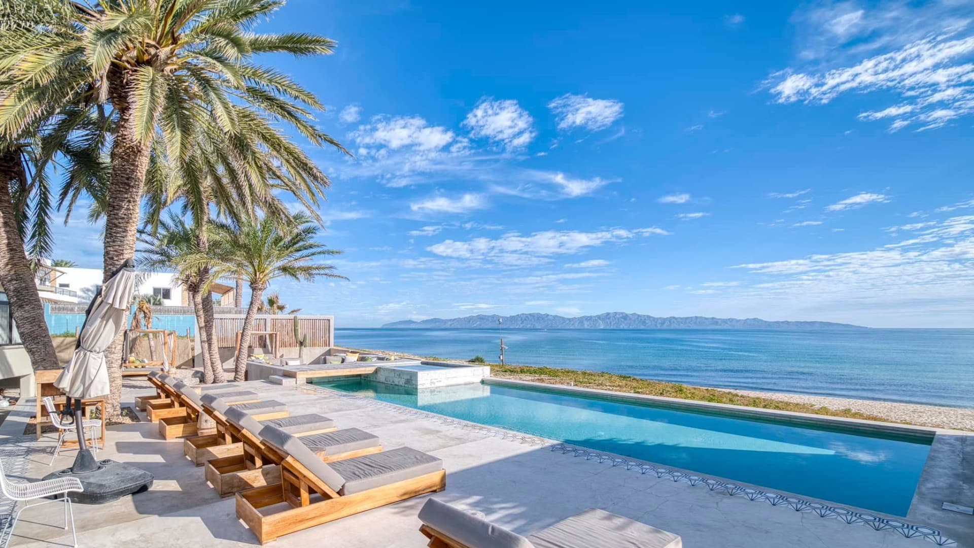 A serene outdoor space featuring a pool, lounge chairs, palm trees, and a beach view under a clear blue sky.