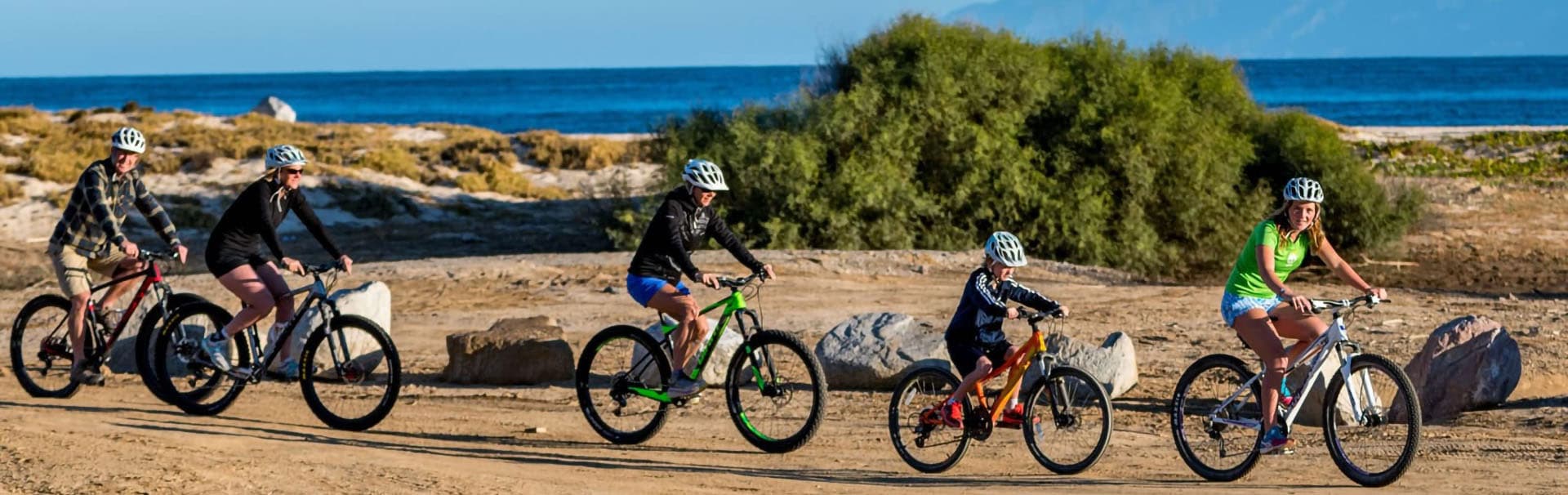 A group of five people riding bicycles along a sandy path with the ocean in the background.