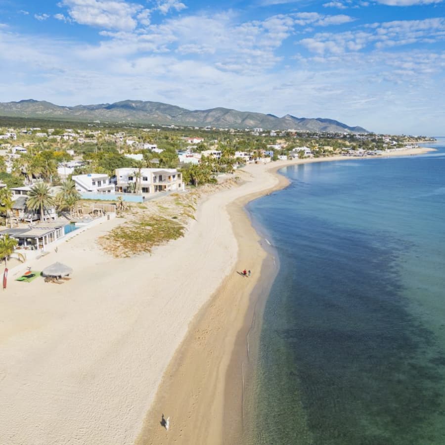 Aerial view of a sandy beach with clear water and distant mountains, lined with palm trees and residential buildings.