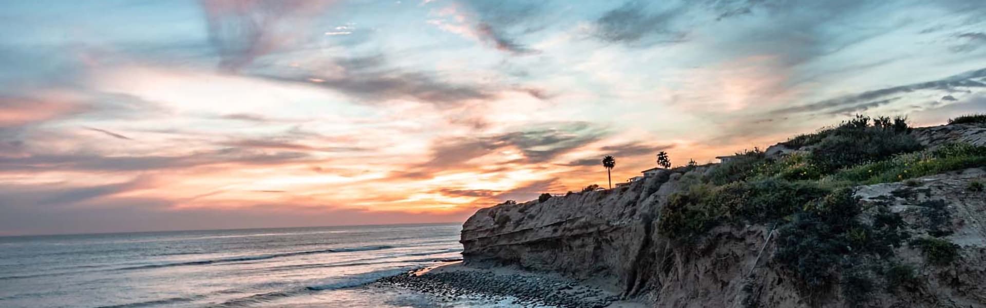 Sunset over a rocky coastline with palm trees silhouetted against colorful clouds.